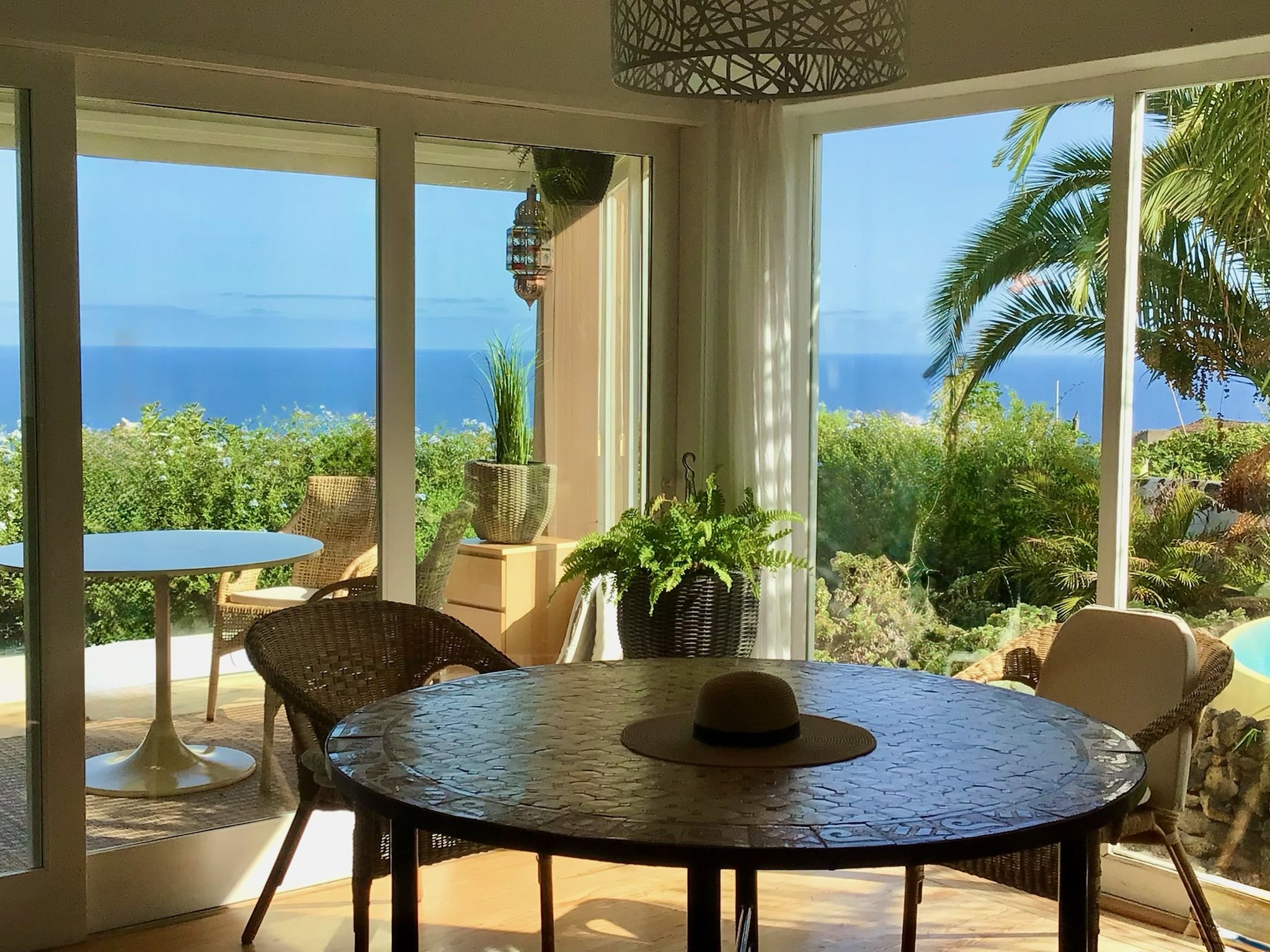 Bellamare: Runder Mosaiktisch mit Rattanstühlen vor großen Fenstern mit Blick über den Garten auf das Meer