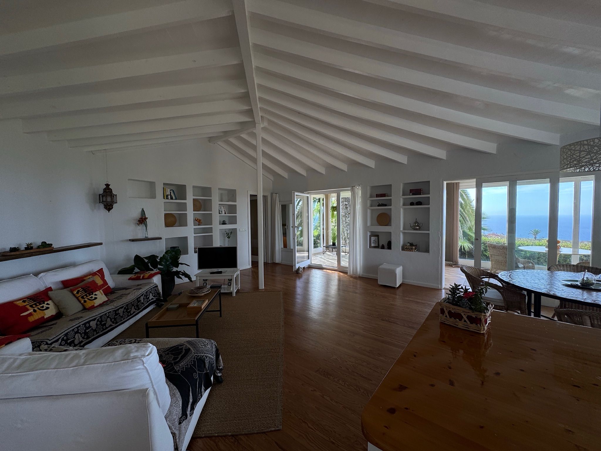 Bellamare: Offener Wohn- und Essbereich mit Sofa, Wandregalen und rundem Esstisch, durch die Fenster Meerblick
