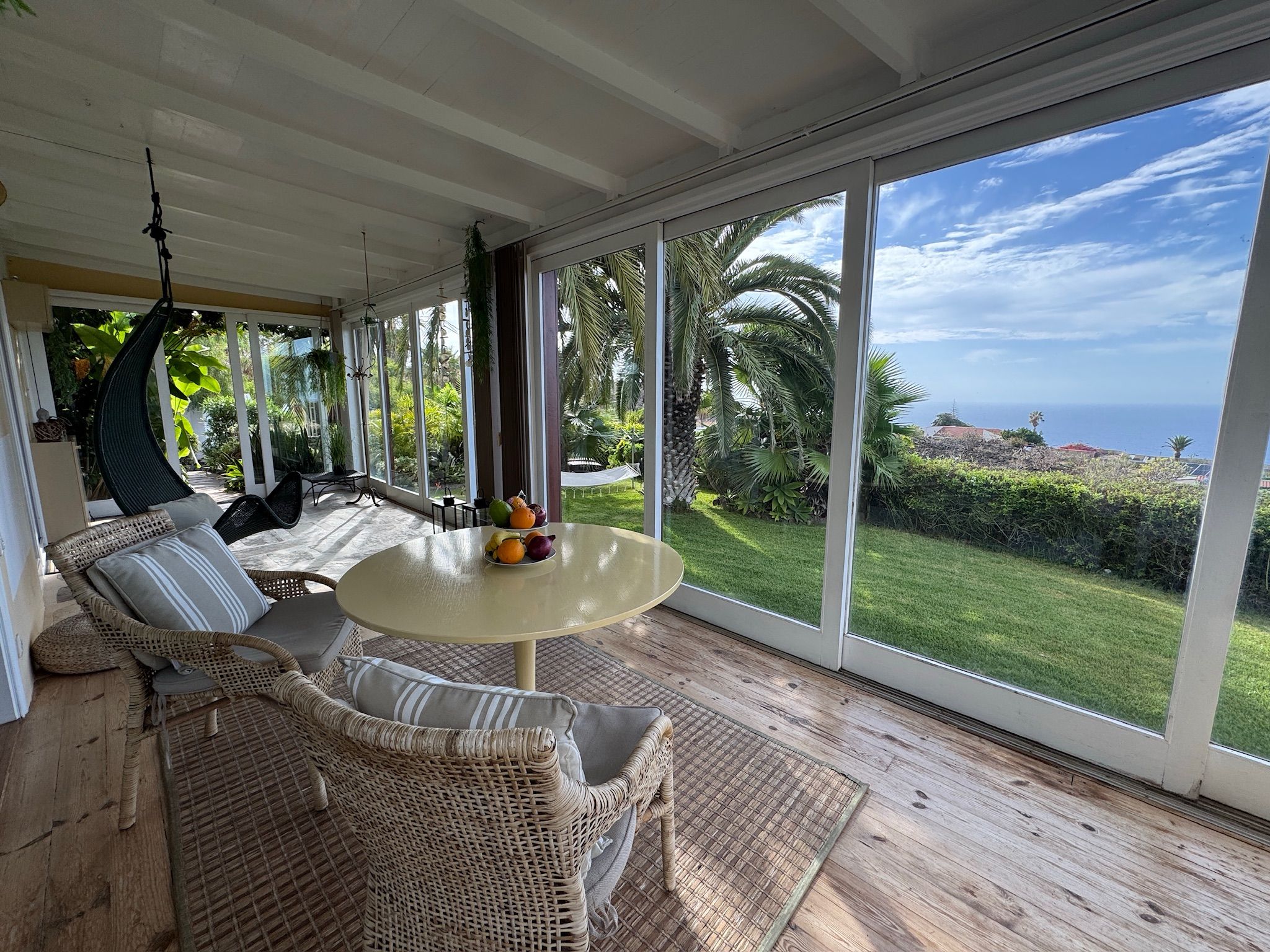 Bellamare: Wintergarten mit Rattansesseln, rundem Tisch und Hängesessel, Blick über den Rasen auf das Meer